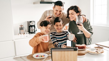 Uma família com quatro elementos (um homem e uma mulher jovens e dois rapazes) reúne-se frente ao ecrã de um tablet, disposto sobre uma mesa onde são visíveis alguns pratos contendo sobremesas. O homem e a mulher seguram copos com vinho, e os rapazes canecas de loiça. Todos erguem os copos ou canecas na direção do ecrã, em jeito de brinde. Atrás, em segundo plano, é visível o balcão e armários brancos de uma cozinha, com alguns pequenos eletrodomésticos e um exaustor nos tons preto e prateado.
